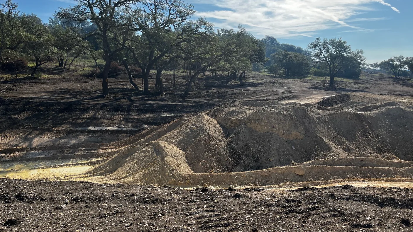 Cleared ground with caliche spoil piles and live oaks under a blue sky
