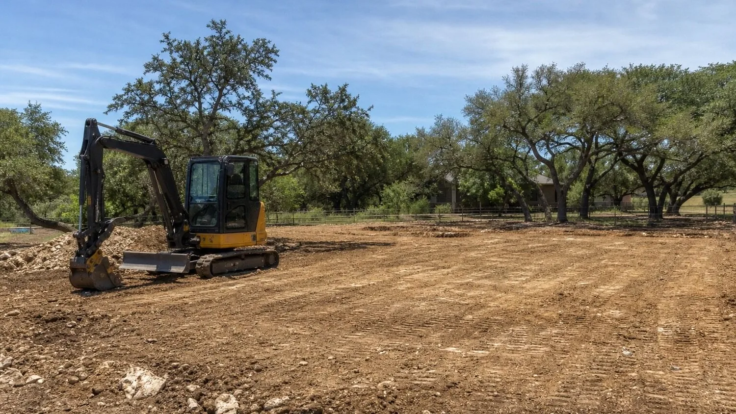 Compact excavator on a cleared lot ringed by live oaks and limestone