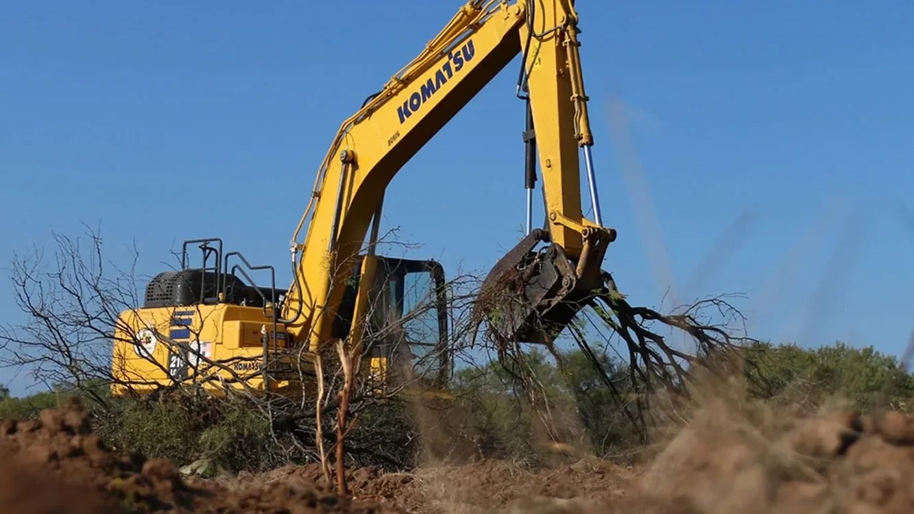 Excavator with a grapple pulling mesquite out of brush country ground