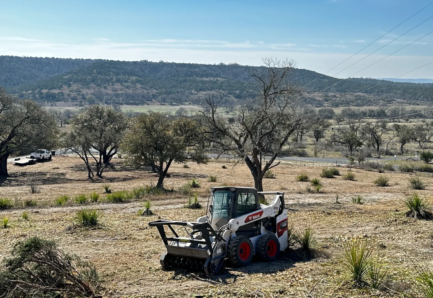 Compact track loader clearing brush on a hill country lot with cedar and live oak