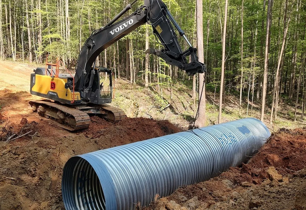 Excavator setting a large metal culvert pipe into a cut