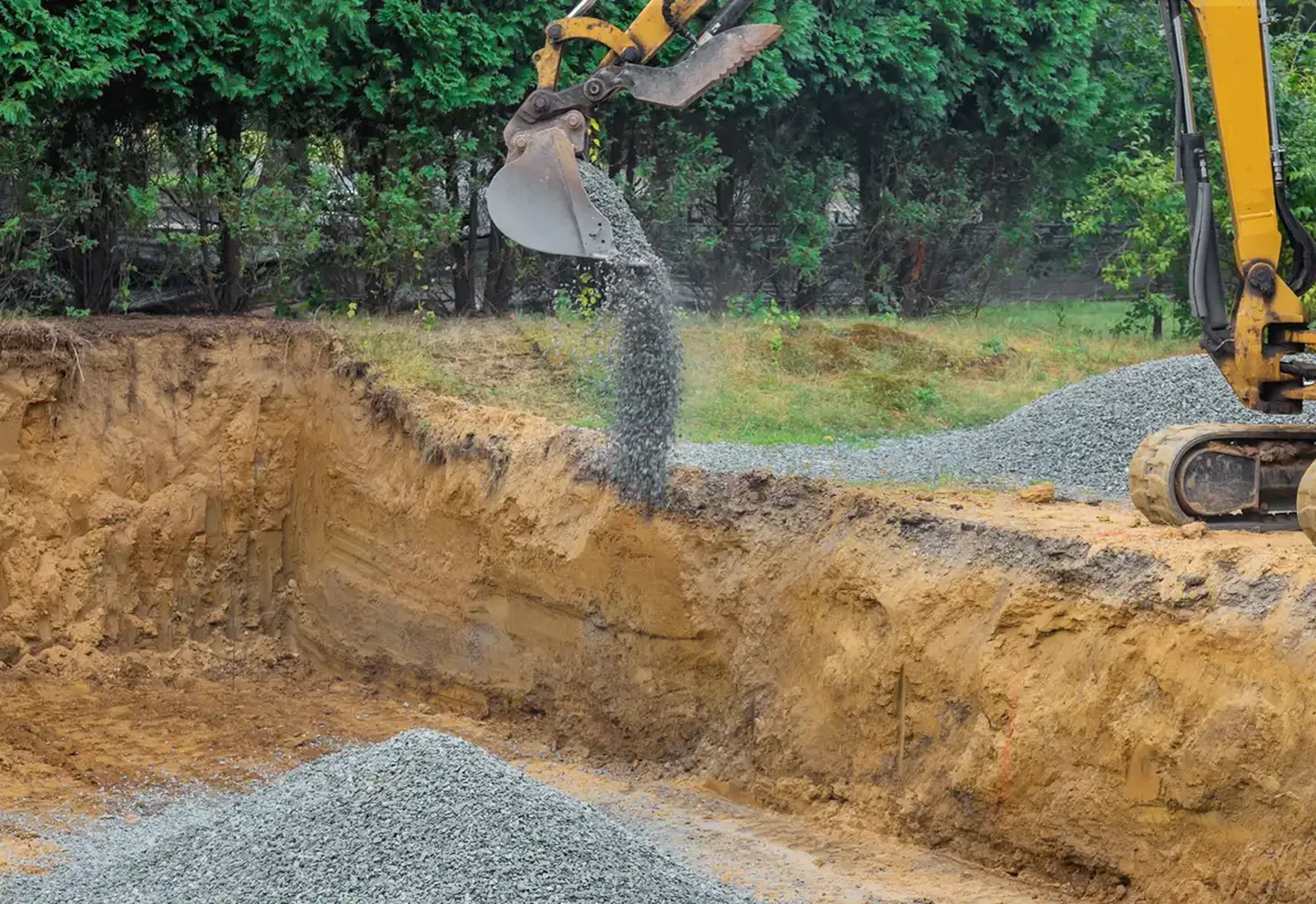 Excavator bucket dropping gravel backfill into an open trench